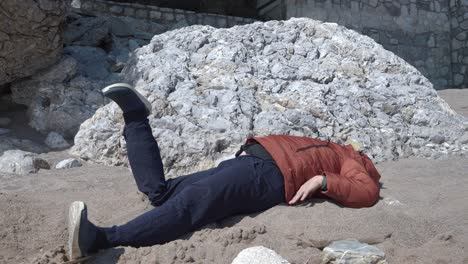 plump man in orange jacket raises torso leaning on arms and looks at broken leg unusually bent near beach rocks on sunny spring day