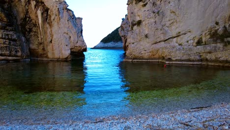 Panning-handheld-shot-of-the-boat-behind-the-rocky-cliffs-in-the-clear-glistering-waters-on-the-beach-Stiniva,-on-the-island-Vis,-Croatia