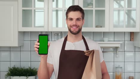 a man holds a smartphone with a green screen in his hand and smiles while standing in the kitchen. home delivery app