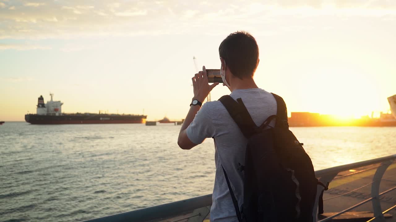 Man With Backpack Taking Photo Of A Ship Moored At Quiet Ocean At The Port Of Puerto Ingeniero White, Buenos Aires, Argentina