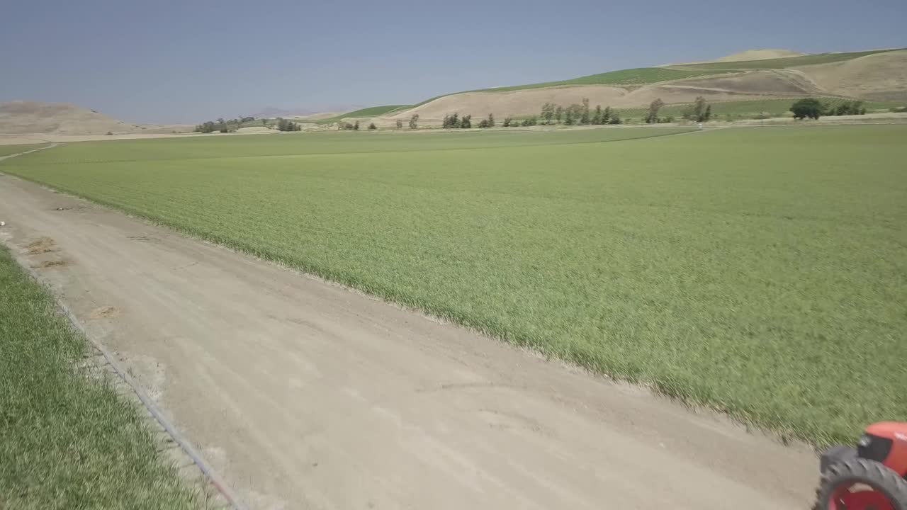tierras de cultivo rurales, imágenes aéreas, moviéndose hacia adelante y hacia arriba sobre tractor rojo, extensos cultivos verdes y colinas doradas y cielo azul en el fondo, 4k 24fps, gira por el camino de campo