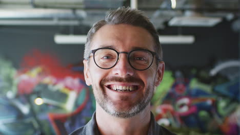 Middle-aged-white-man-smiling-to-camera-in-front-of-mural-in-creative-workplace,-close-up-head-shot