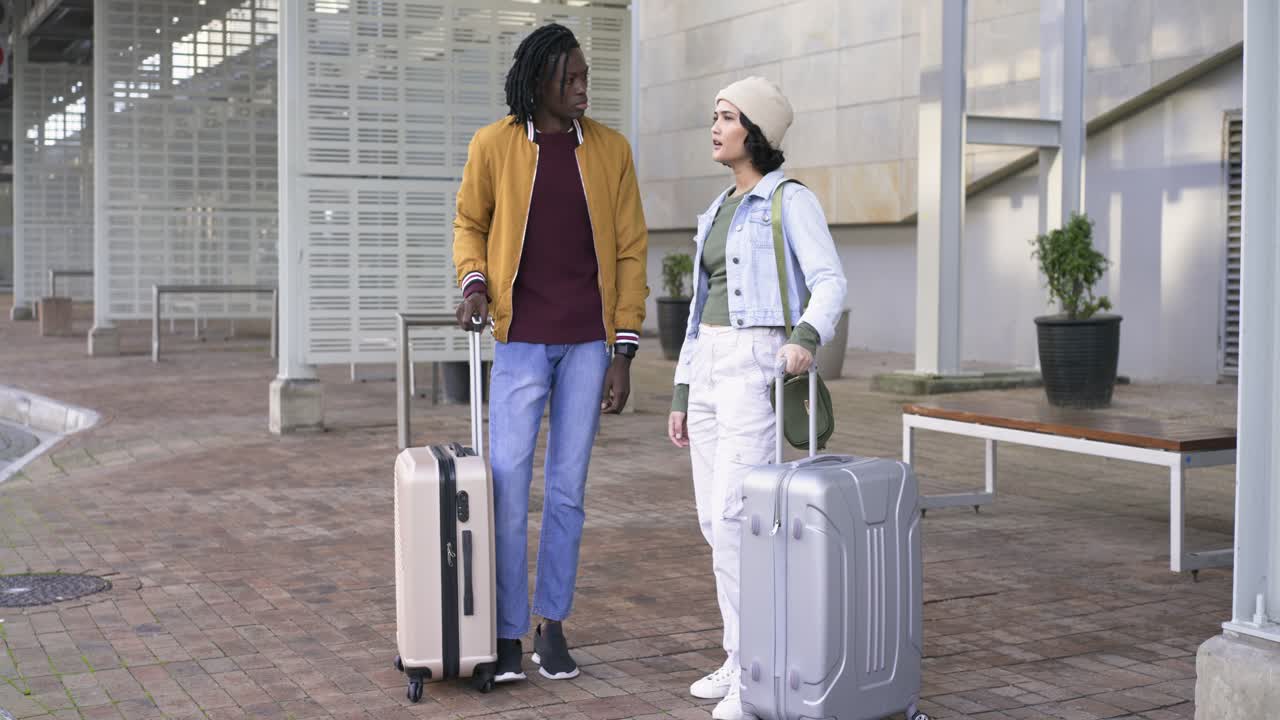 Diverse couple arriving at courtyard pulling suitcases, chatting then raising smartphone for selfie