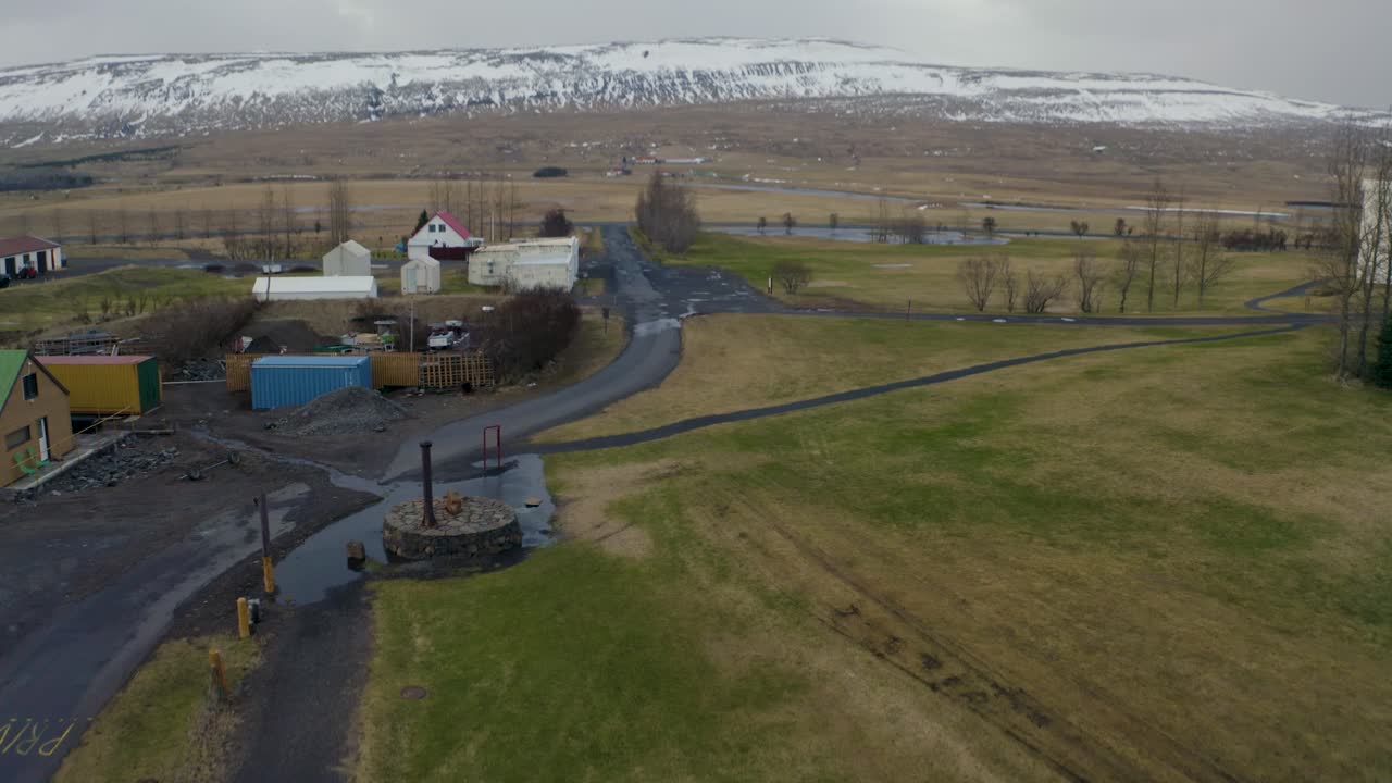 Drone footage showcasing a small Icelandic village surrounded by green fields, scattered houses, and a snow-capped mountain range in the background. A serene countryside scene in Iceland.