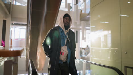 young man in hoodie and jacket standing by escalator in modern shopping mall, holding onto railing, observing his surroundings. bright atmosphere with festive decor, urban interior, casual style