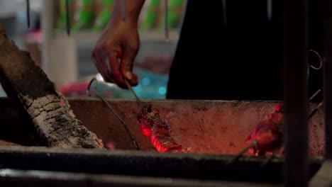 a man’s hand turning meat kabob skewers on an open flame grill in the outdoors