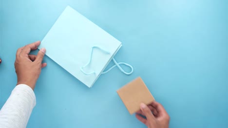 manos colocando una caja de regalos en una bolsa de compras azul claro en una superficie azul claro.