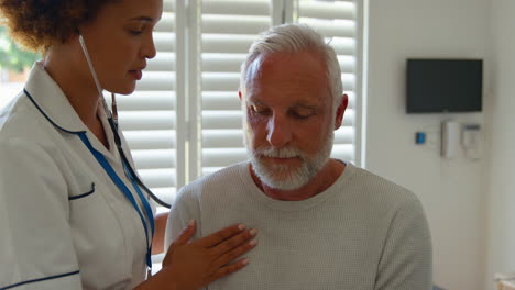 female nurse wearing uniform listening to senior male patient's chest in private hospital room