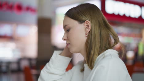 side profile of a woman sitting in a mall with thoughtful expression, hand brushing her hair, surrounded by blurred vibrant decor, soft lighting, and a busy background