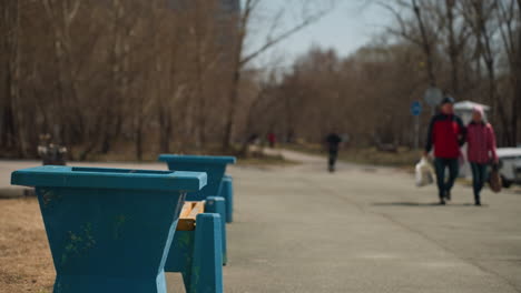 close view of blue park benches in the foreground with people strolling in the background, with a blur view of someone on black clothing riding an electric scooter