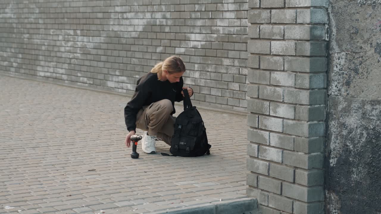 Uomo bianco cammina lungo un muro di mattoni con zaino e caffè, indossa pantaloni cargo casual e scarpe da ginnastica bianche, passo deciso su un marciapiede urbano, energia da pendolare, luce mattutina nuvolosa, città autentica.