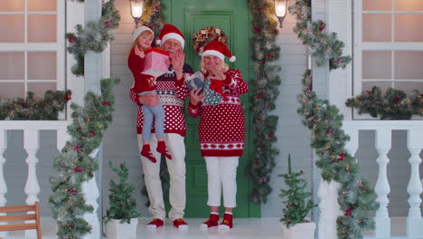 Senior-grandmother-grandfather-with-granddaughter-standing-at-Christmas-house-porch-waving-hello-hi