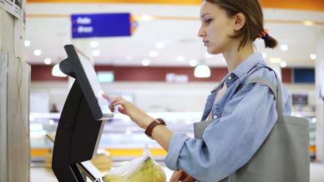 una niña pesa plátanos en una balanza electrónica en un supermercado y pone una etiqueta adhesiva con un precio. plátanos en un paquete de celofán. vista lateral
