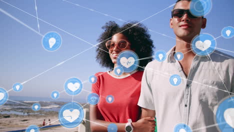 red de iconos digitales contra una pareja biracial tomándose de la mano caminando por la playa