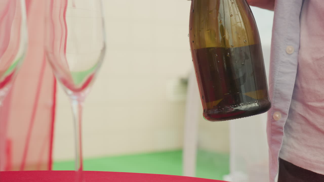 Appuntamento su un tetto: uomo bianco che appoggia una bottiglia di champagne su una tovaglia rossa accanto a due calici da vino, goccioline sulla bottiglia, manica di una camicia casual visibile, luce diurna soffusa, atmosfera intima e celebrativa, tintinnio leggero