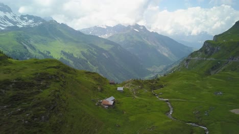 Aerial-dolly-in-flying-over-small-cottages-in-green-valley-hillside,-verdant-mountains-and-forest-in-background-on-a-cloudy-day,-Switzerland-Alps