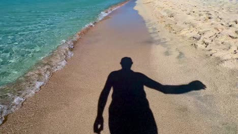 silhouette shadow of man putting on sunglasses and walking on sandy beach along turquoise sea water shoreline