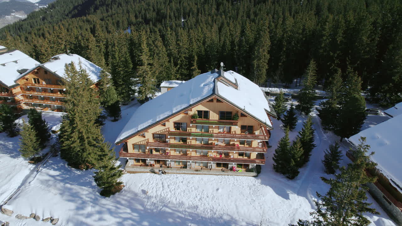 Une vue aérienne plongeante sur un chalet de ski en bois confortable dans la magnifique station de Méribel enneigée, avec des skieurs et des snowboarders passant entre les pins lors de leurs luxueuses vacances d’hiver dans les Alpes françaises.