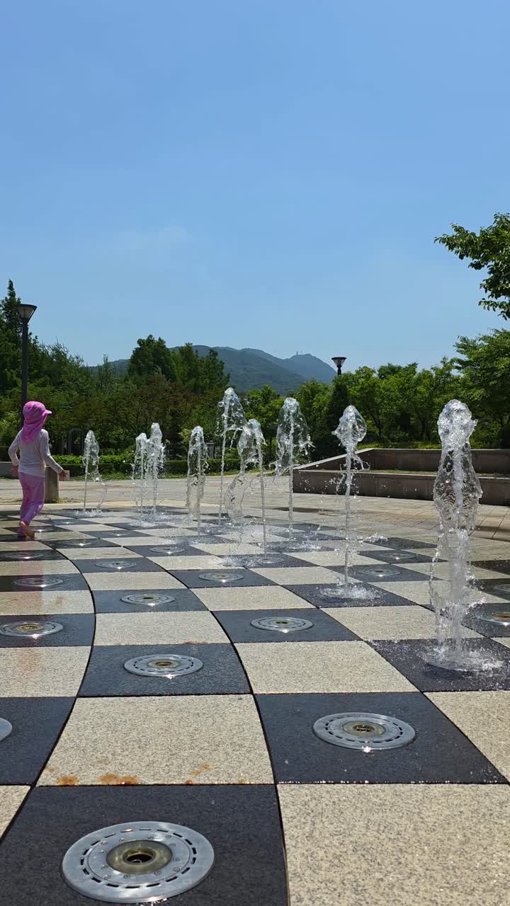 A joyful little girl in pink swimwear and sun hat playfully kicks water jets at a ground fountain in Seocho Park, Seoul, enjoying a sunny summer day outdoors
