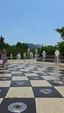 a joyful little girl in pink swimwear and sun hat playfully kicks water jets at a ground fountain in seocho park, seoul, enjoying a sunny summer day outdoors