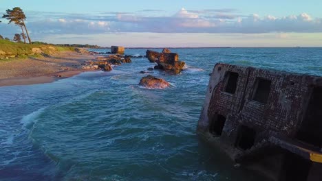 vista aérea de los edificios de fortificación costeros abandonados en los fuertes del norte de karosta en la playa del mar báltico, olas salpicadas, hora dorada puesta de sol, gran tiro de drones avanzando