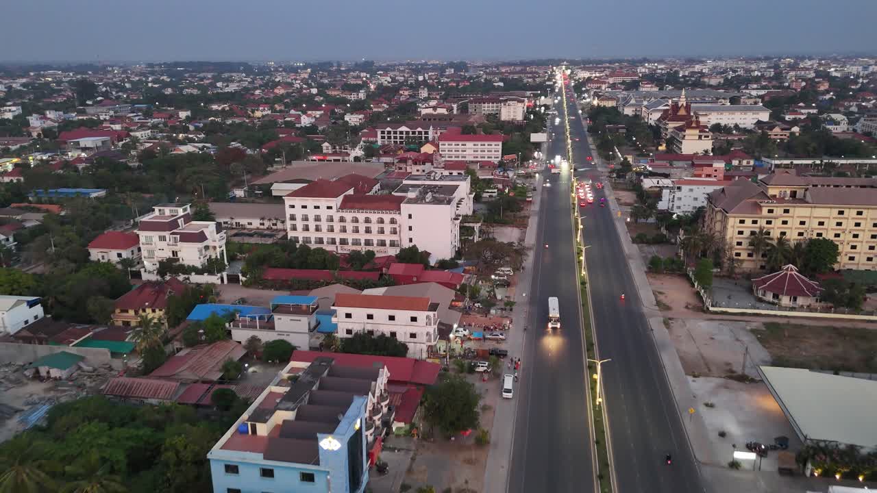 An elevated, wide shot showcasing a bustling urban environment at dusk or dawn. A long, well-lit multi-lane road stretches through the center of the frame