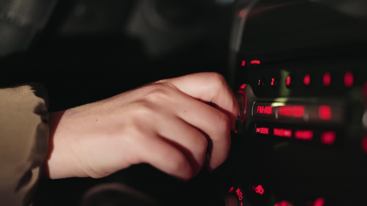 vista en primera persona de la mano de una mujer girando el botón de la radio fm dentro de un coche por la noche. el tablero de instrumentos está iluminado con luces rojas