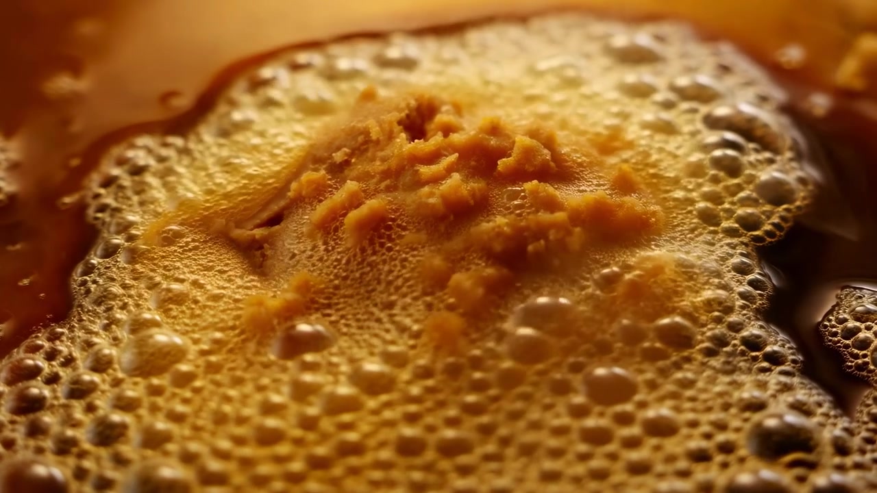 Vue macro rapprochée montrant des fragments écrasés orange se dispersant dans un liquide ambré brillant, tandis que la mousse se répand, des bulles montent et le matériau texturé crée une couleur tactile riche et du mouvement