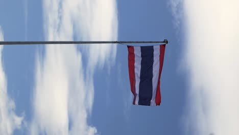 footage in portrait orientation of thailand national flag hoisted on a white tall steel pole blown by the wind creating an animated wave on a backdrop of a clear blue sky and white clouds