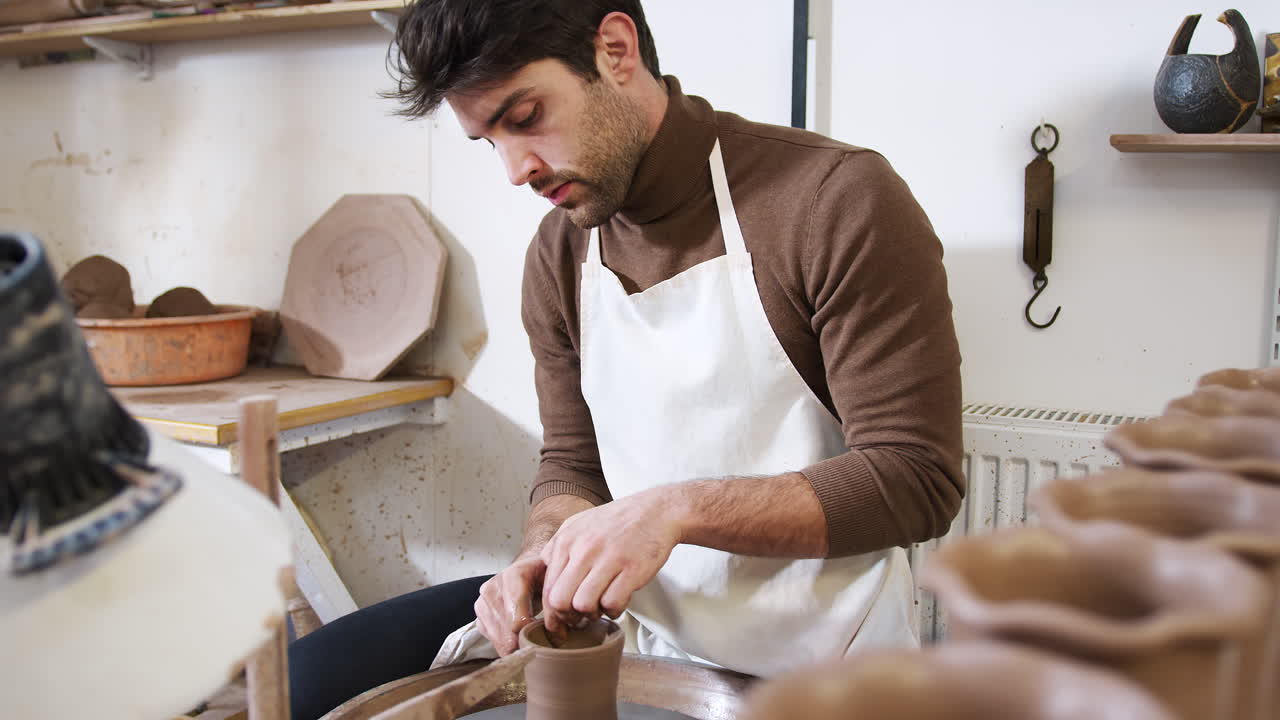 Young Man Wearing Apron Working At Pottery Wheel In Ceramics Studio