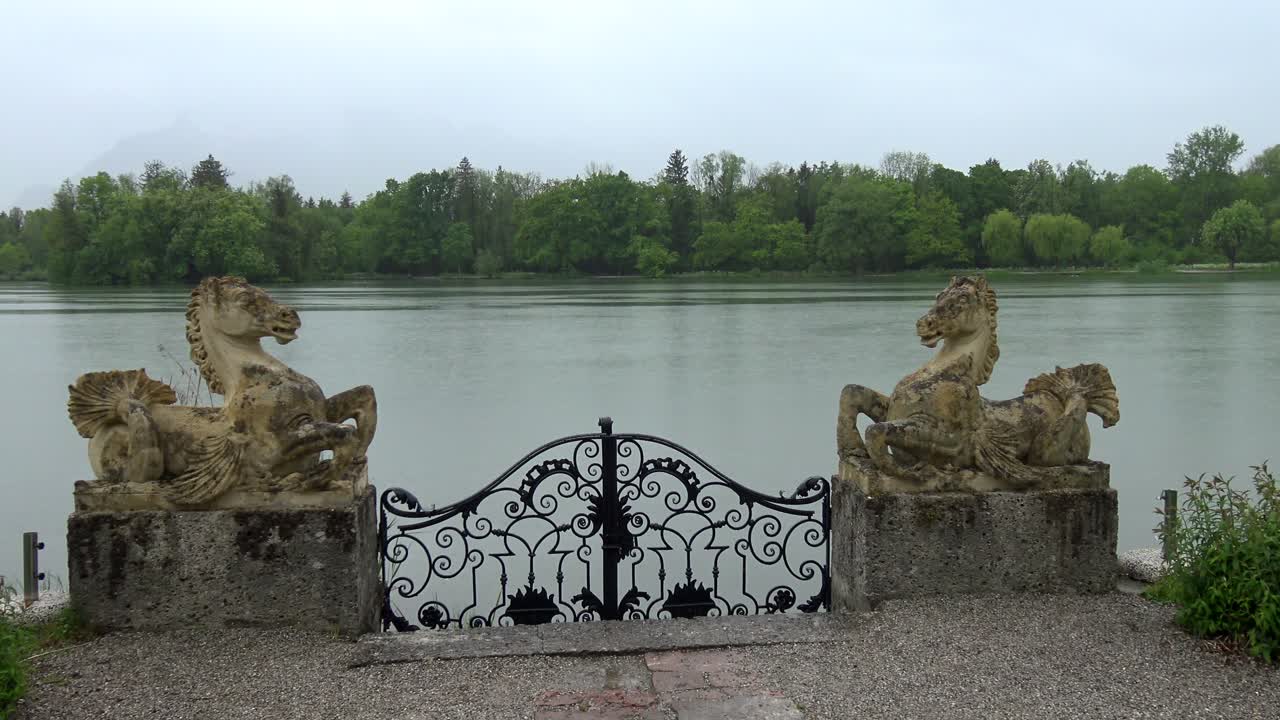 Two ancient stone horse sculptures flank an iron gate leading down to a lake in the rain.