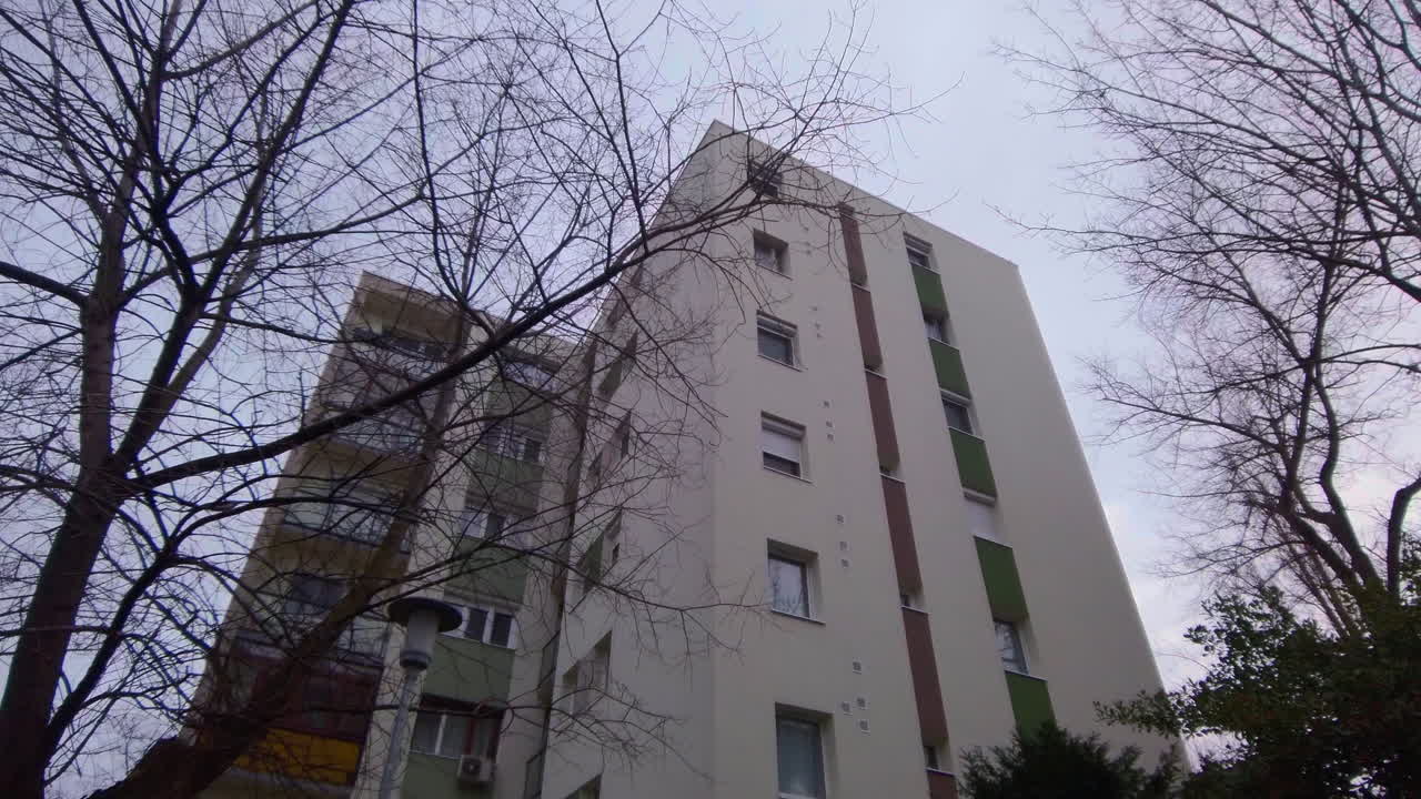 Et førstepersonsperspektiv der går fremad for at afsløre en høj beboelsesejendom på Ecseri ut i Budapest. Den har brune altaner og nøgne vintertræer under en blid overskyet himmel i februar 2026.