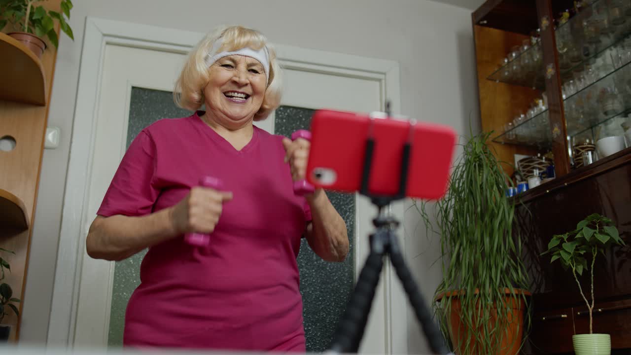 Senior elderly old woman watching online distance workout exercises with dumbbells on mobile phone