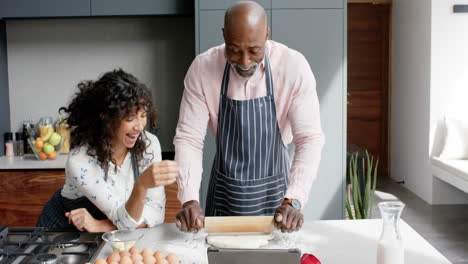 Happy-biracial-couple-wearing-aprons-baking-and-using-tablet-in-kitchen,-slow-motion