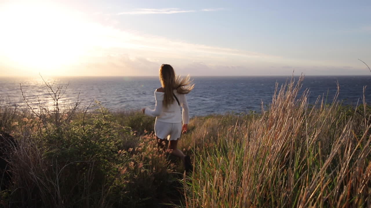 una mujer vestida de blanco camina a través de la hierba alta con vistas a una hermosa vista al mar al atardecer