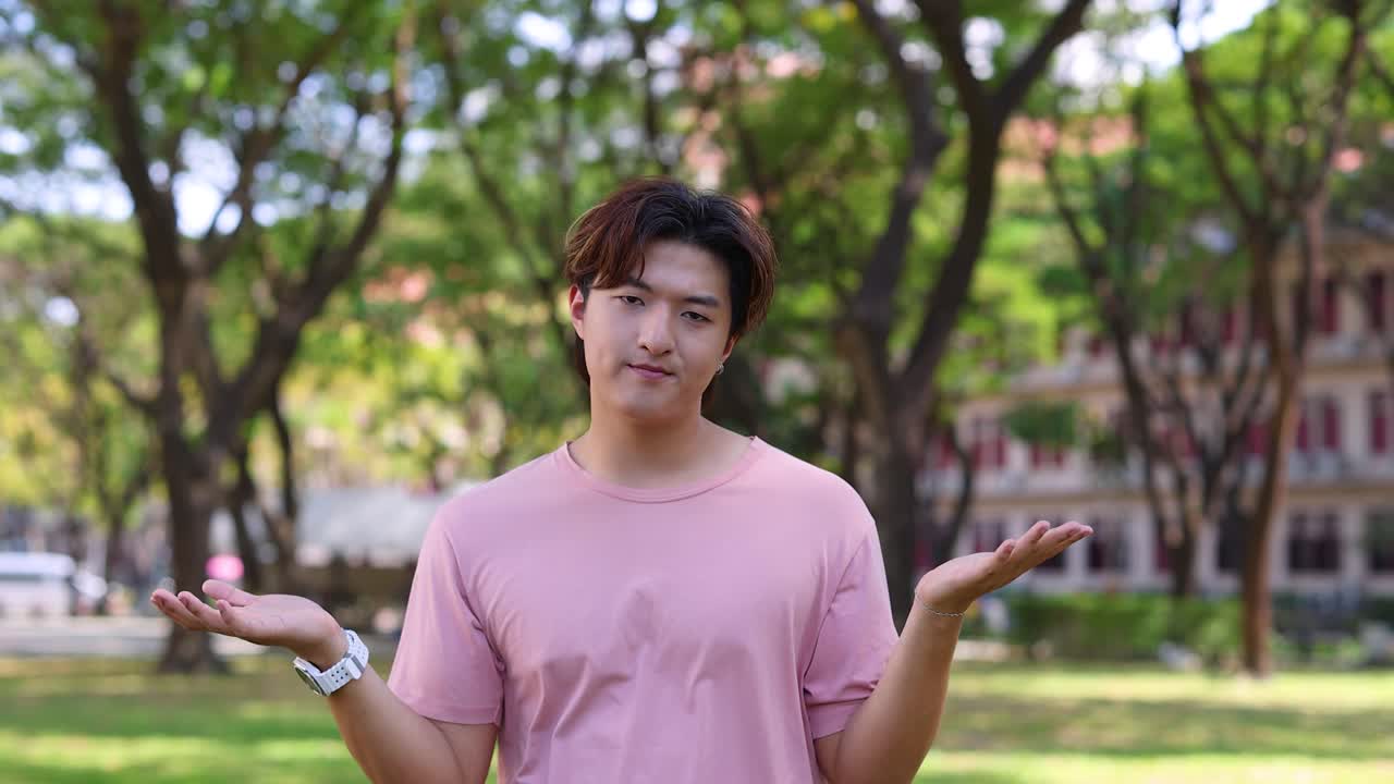 Một thanh niên châu Á mặc áo sơ mi hồng nhún vai bối rối trong công viên đầy nắng.