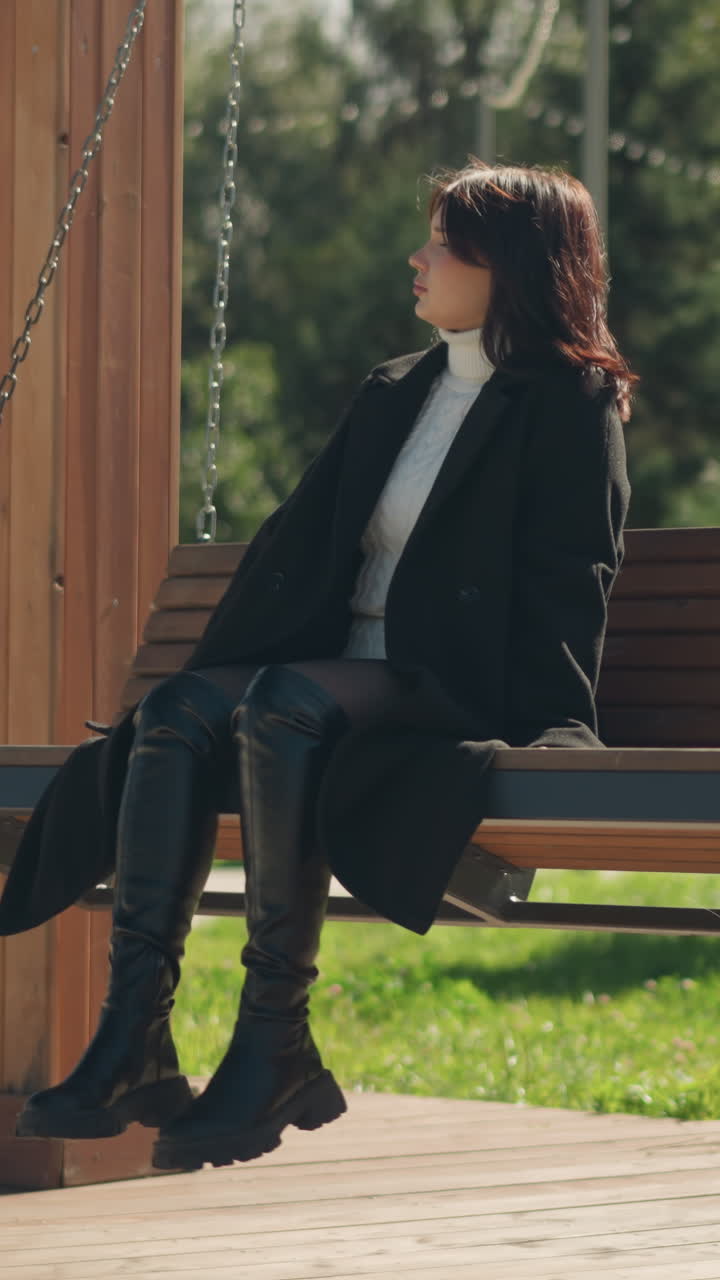 검은 코트와 가죽 부츠를 입은 여인이 공원 안의 나무 스 의자에 앉아 푸른 공원 풍경과 나무와 꽃의 흐릿한 배경으로 밝은 빛을 즐기고 있습니다.