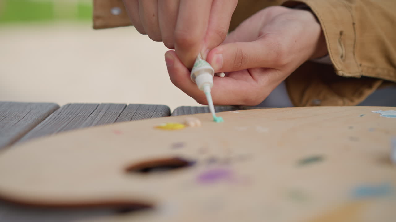 Artist painting outdoors. Painting scene with tactile details. Closeup of artist applying paint outdoors. Outdoor artist concentrates while squeezing paint onto palette with casual attire
