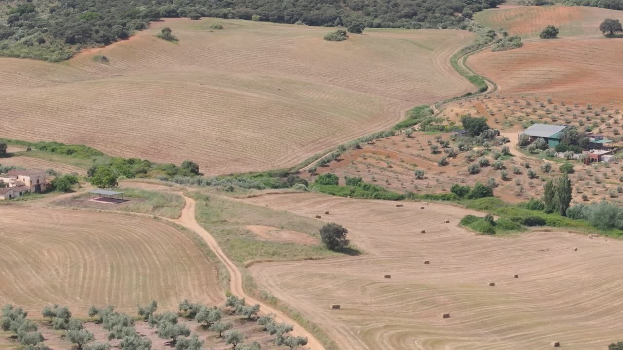 Reverse drone shot at 70mm shows harvested and plowed plots with hay bales in varied earth tones, passing by farms, olive groves and a dirt road leading to a small oak forest