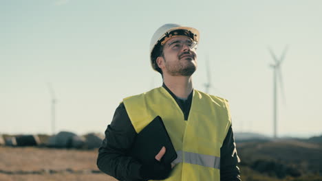 a professional engineer in a field of wind turbines, dressed in safety gear, takes a break to smile at the camera