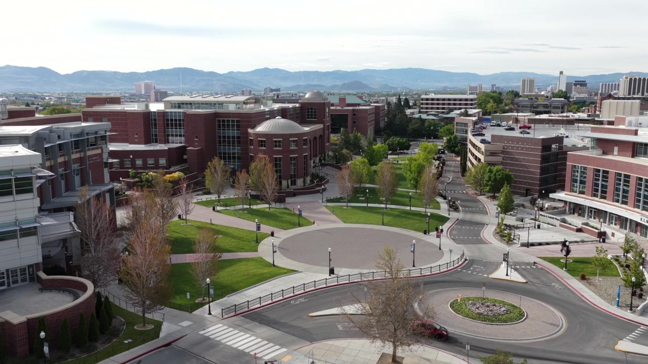 Campus universitario con edifici in mattoni rossi, piazza circolare, rotonda e viali alberati che si estendono verso lo skyline del centro e una catena montuosa sotto un cielo parzialmente soleggiato. Nevada, USA. Approccio aereo.