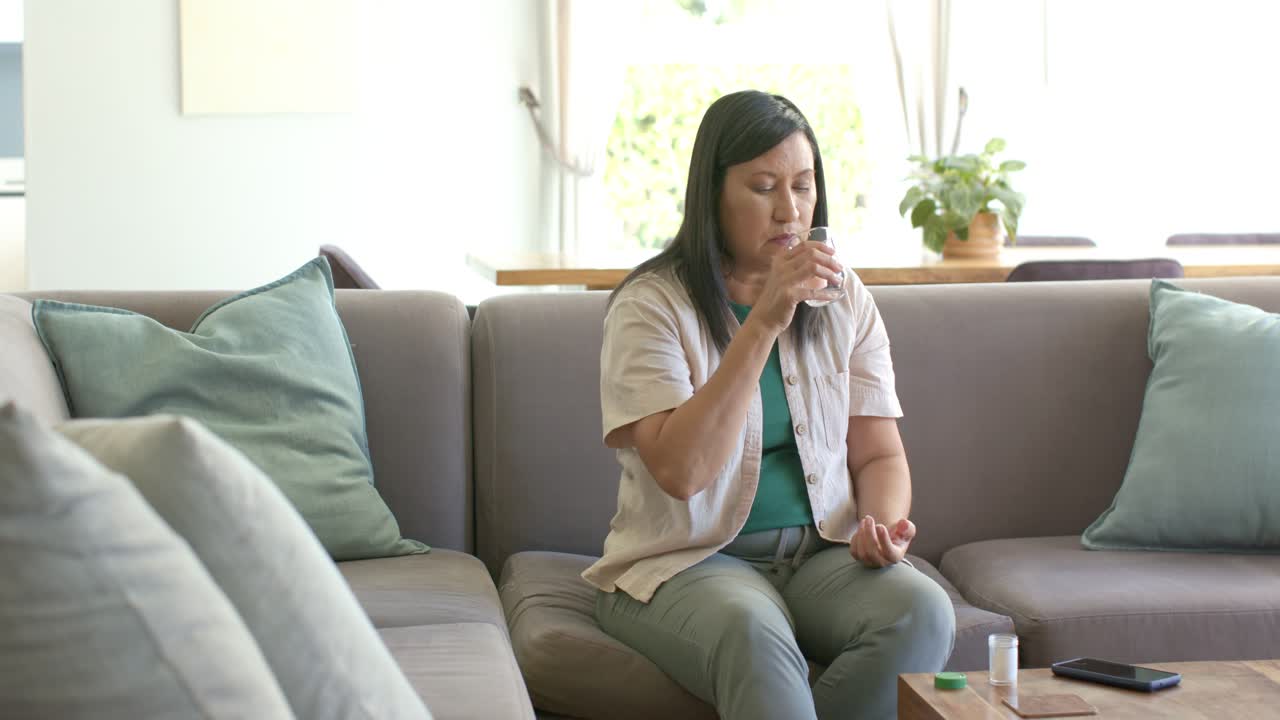 Donna asiatica che esamina una confezione di pillole, versa una pillola e beve acqua sul divano per prendere la medicina