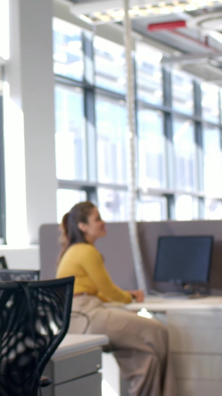 Video verticale: Donna con top color senape si gira, sorridendo alla scrivania mentre un collega entra da sinistra in ufficio