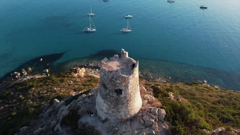 Birds-eye-view-of-Torre-di-Porto-Giunco-tower-standing-tall-on-island-surrounded-by-sea-with-yachts-sailing-and-people-exploring-and-taking-pictures-in-Italy