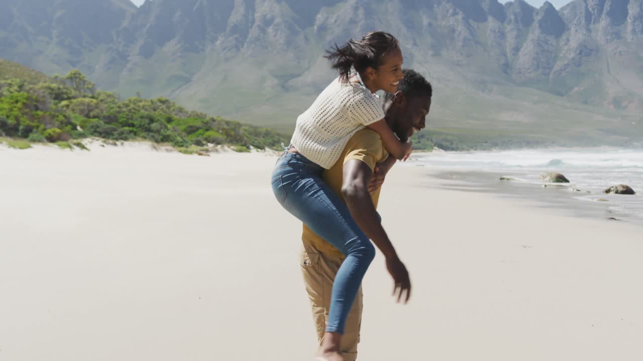 African american man giving piggyback ride to his wife at the beach