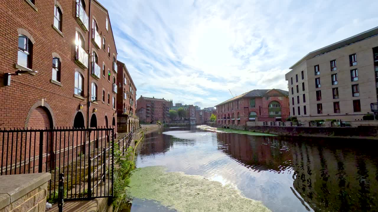 Camera pans alongside a canal bordered by red brick and modern buildings under bright daylight, capturing reflections, water, and urban architecture in Leeds