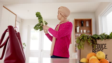 Feliz-Mujer-Birracial-En-Hijab-Con-Compras-En-La-Cocina-De-Casa-Con-Espacio-Para-Copiar,-Cámara-Lenta