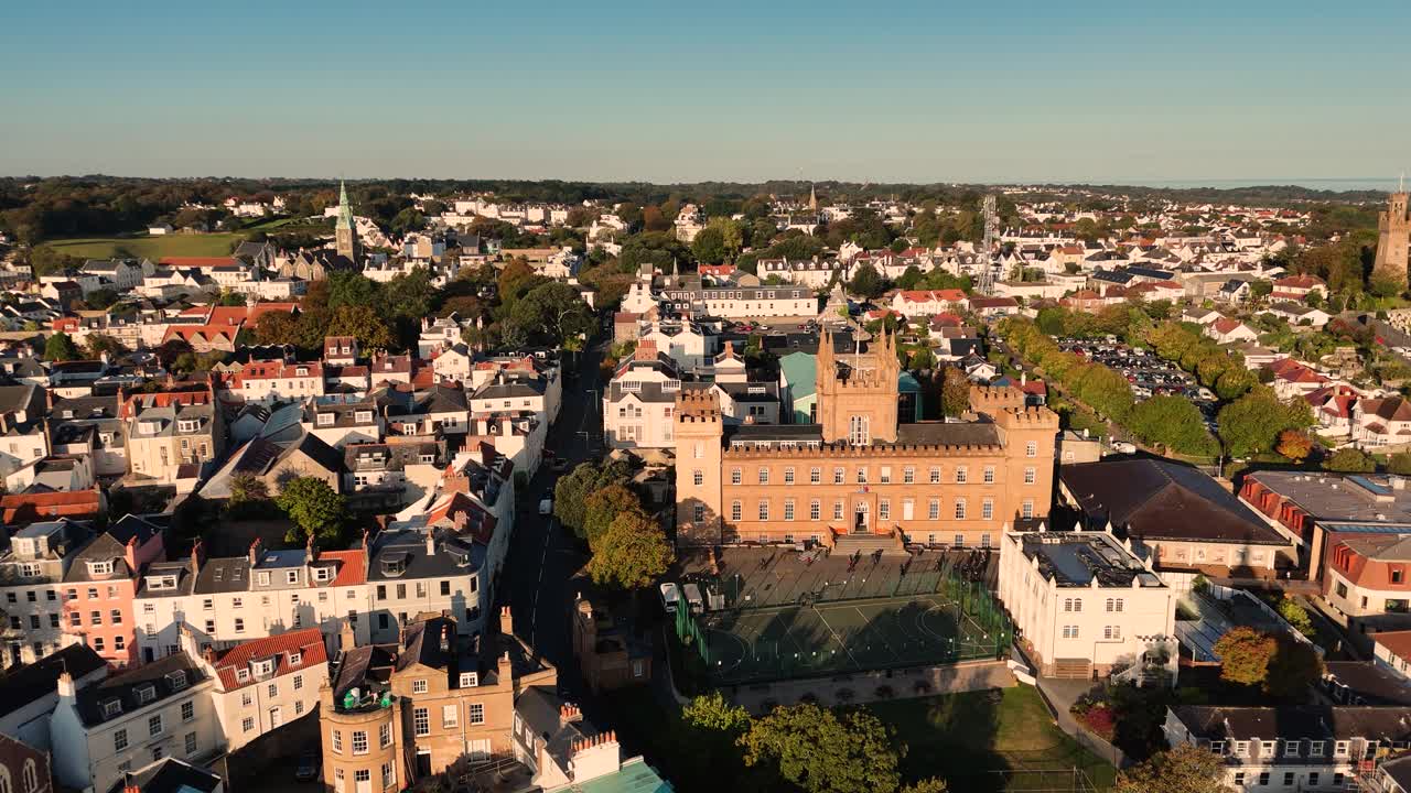 Sakte droneopptak av St. Peter Port, Guernsey, over St. James og hustakene mot Elizabeth College i gyllent solskinn, med utsikt over silhuetten og hele øya på en skyfri sommerdag.