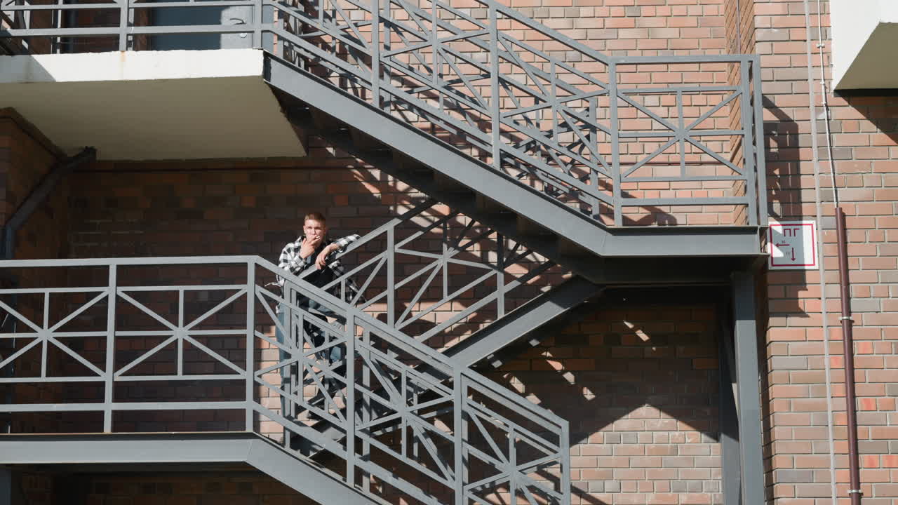 Man in black and white checkered shirt leans casually on metal railing while smoking cigarette on outdoor walkway of multi-level brick building with geometric staircase shadows and industrial design