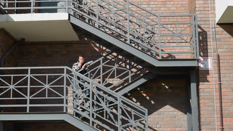 man in black and white checkered shirt leans casually on metal railing while smoking cigarette on outdoor walkway of multi-level brick building with geometric staircase shadows and industrial design